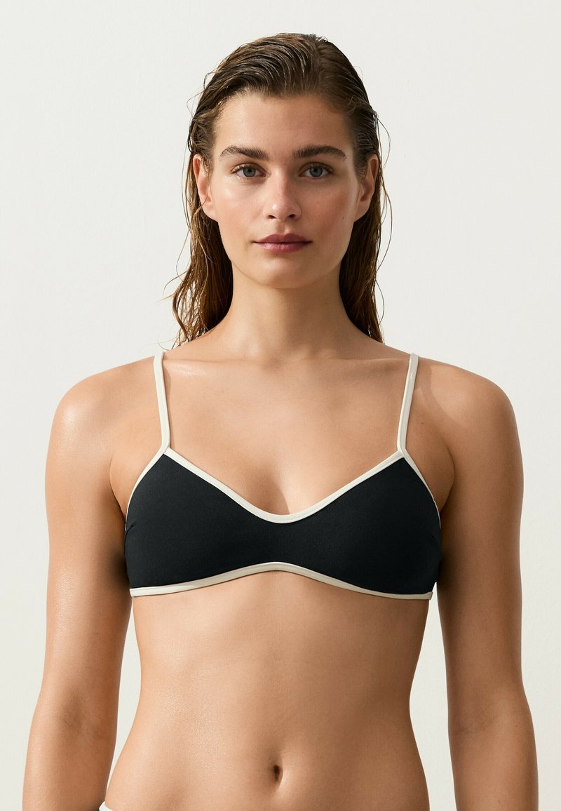 Femme aux cheveux mouillés portant un bralette noir avec une bordure blanche, face à un fond clair uni.
