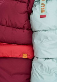 Közelkép bordó és világoskék steppelt dzsekikről, narancssárga és piros címkékkel, melyeken a szövet textúrája és a varrásvonalak láthatók.