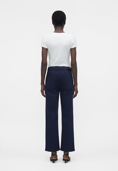 Άτομο με λευκό κοντομάνικο μπλουζάκι, σκούρο μπλε φαρδιά παντελόνια και μαύρα πέδιλα με τακούνι, στέκεται γυρισμένο προς τα πίσω σε λευκό φόντο.