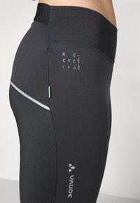 Fekete sportleggings újrahasznosított anyagból. Reflektorcsíkkal, zöld kiegészítővel és oldalán nyomtatott logóval rendelkezik.