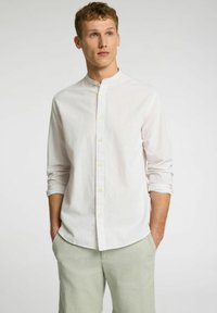 Camisa blanca a rayas con cuello de banda, de mangas largas enrolladas en el puño, combinada con pantalones de lino verde claro.