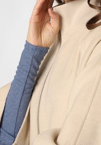 Beige lynlåsejakke med høj krave, lavet af et blødt, struktureret stof. Blå ribbede lange ærmer nedenunder, med et diskret mønster.