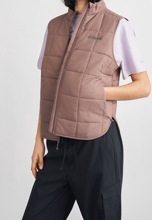 Person, die eine mauve gesteppte ärmellose Weste über einem weißen kurzärmeligen Hemd und schwarzen Hosen trägt, mit einer Hand in der Westentasche.