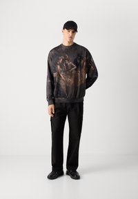 Mandlig model iført sort cap, mørk sweatshirt med klassisk maleri-tryk, sorte cargo-bukser og sorte skinnende sko, stående mod hvid baggrund.
