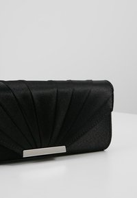 Pochette en satin noir avec un design en éventail plissé et une fermeture rectangulaire argentée, posée sur une surface blanche devant un fond gris clair.