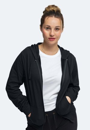 Schwarze Kapuzenjacke mit einem Reißverschluss, Fronttaschen und gerippten Bündchen. Darunter ein weißes Shirt. Glatte Textur, lässiges Design.