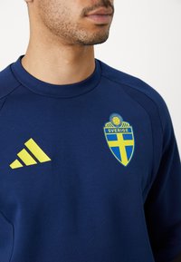 Temno modra majica s kapuco in okroglo ovratnikom, ima rumen logotip Adidas in nacionalni grb Švedske v modro-rumeni barvni shemi.