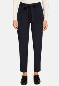 Pantalon bleu marine fuselé en tissu lisse avec une taille nouée à l'avant et deux poches latérales, porté avec des mocassins noirs.