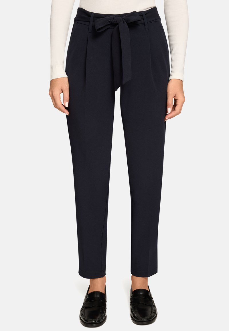 Pantalon bleu marine fuselé en tissu lisse avec une taille nouée à l'avant et deux poches latérales, porté avec des mocassins noirs.