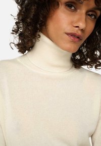 Cremfarbener Rollkragenpullover aus weichem Rippstrickstoff, mit hohem Kragen und glatter Textur, der eng am Hals anliegt.