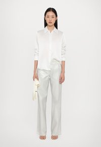 Witte satijnen overhemd met lange mouwen, gecombineerd met glanzende zilveren wijde broek. Model houdt een lichtgekleurde trui vast.