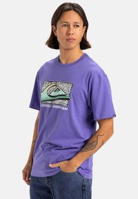 Camiseta de manga corta morada hecha de algodón, con un diseño gráfico de olas y montañas en verde y negro en la parte frontal.