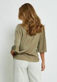 Olive green silk blouse with three-quarter sleeves, soft sheen, subtle drape, and a rounded hem. Styled with light-colored trousers.
