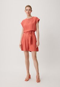 Korallenbluse mit kurzen Ärmeln und Schlitzdetails, kombiniert mit hochtaillierten Korallenshorts mit einem gebundenen Gürtel, ergänzt durch Riemchensandalen mit Blockabsatz.