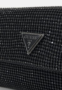 Černá kabelka na spojení pokrytá malými kulatými černými kamínky. Obsahuje trojúhelníkové stříbrné logo s nápisem "GUESS" a otazníkem. Hladký povrch.