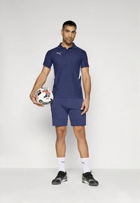 Sötétkék galléros póló fehér díszítéssel, hozzáillő rövidnadrág és fekete sportcipő. A modell focilabdát tart, bemutatva a sportos öltözéket.