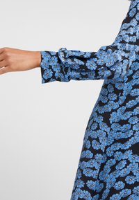 Sininen kukkakuviollinen mekko mustalla taustalla, jossa on pitkissä hihoissa röyhelövärjäys ja sileä, kevyt kangas.