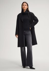Cappotto nero con design strutturato, maniche lunghe e colletto a revers, abbinato a un dolcevita nero e jeans a zampa scuri. Stivali neri con tacco.