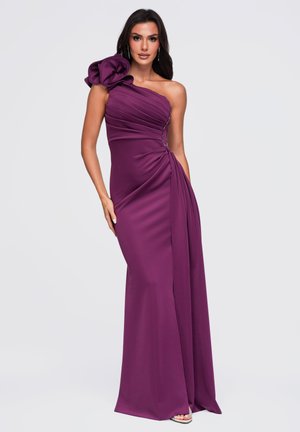 Une femme présente une robe de soirée prune longue jusqu’au sol, avec une encolure asymétrique dégageant une épaule, un détail floral sculptural et une jupe drapée.
