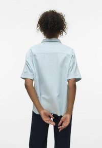 Camicetta azzurra chiara a maniche corte con colletto, caratterizzata da una vestibilità rilassata e dettagli con bottoni sulle maniche. Pantaloni in denim visibili sotto la camicia.