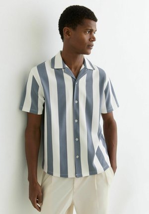 Mężczyzna w koszuli na krótki rękaw z szerokimi pionowymi szarymi i białymi paskami, stojący z rękami w kieszeniach kremowych spodni, patrzący w prawo.