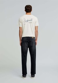 Tricou alb cu mânecă scurtă și text albastru pe spate, asortat cu blugi închisi la culoare. Îmbrăcămintea are o croială lejeră și un stil casual.
