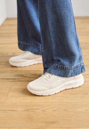 Białe siatkowe sneakersy o teksturowanym wykończeniu, z grubą podeszwą i subtelnym logo. Połączone z dżinsami o szerokich nogawkach na drewnianej podłodze.