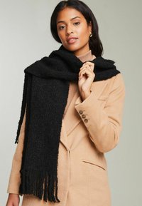 Sort tekstureret tørklæde med frynsede kanter, båret over en beige frakke. Materialet af tørklædet ser blødt og varmt ud, hvilket supplerer farverne i outfittet.