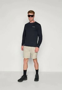 Majica dugih rukava u tamnoplavoj boji s logom, uparena s bež kratkim hlačama. Crne čizme i sunčane naočale upotpunjuju odjevnu kombinaciju. Jednostavan dizajn, funkcionalna odjeća.