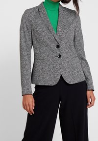 Blazer à motif chevrons noir et blanc avec deux boutons, revers crantés et coupe slim. Associé à un col roulé vert en dessous.