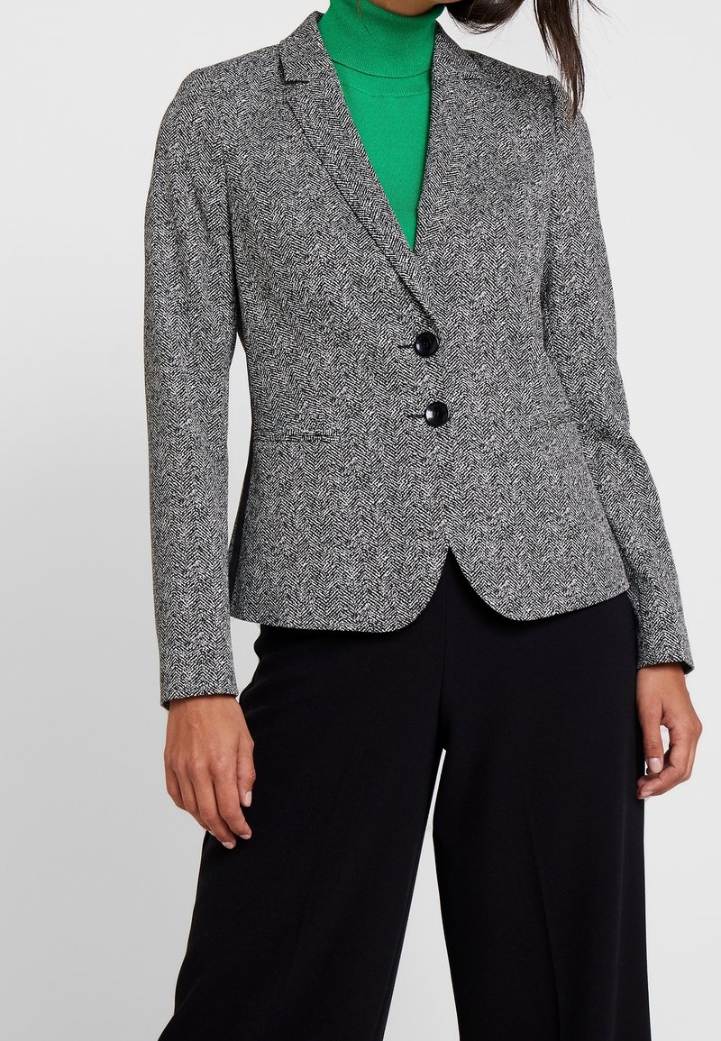 Blazer à motif chevrons noir et blanc avec deux boutons, revers crantés et coupe slim. Associé à un col roulé vert en dessous.