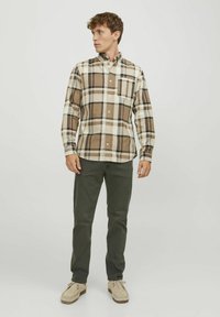 Hombre con una camisa de cuadros marrón y crema, pantalones verdes oscuros y zapatos beige, de pie contra un fondo claro liso, mirando hacia la izquierda.