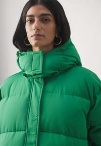 Žalia pūkinė striukė su aukštu kaklu, turinti siuvinėtą tekstūrą, užtrauktukus ir tvirtą prigludimą, sukurta šilumai ir patogumui.