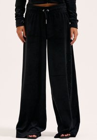 Zwarte wijde pantalon van zacht, fluwelig materiaal. Heeft een elastische tailleband en een zijdetail met kristalversiering.