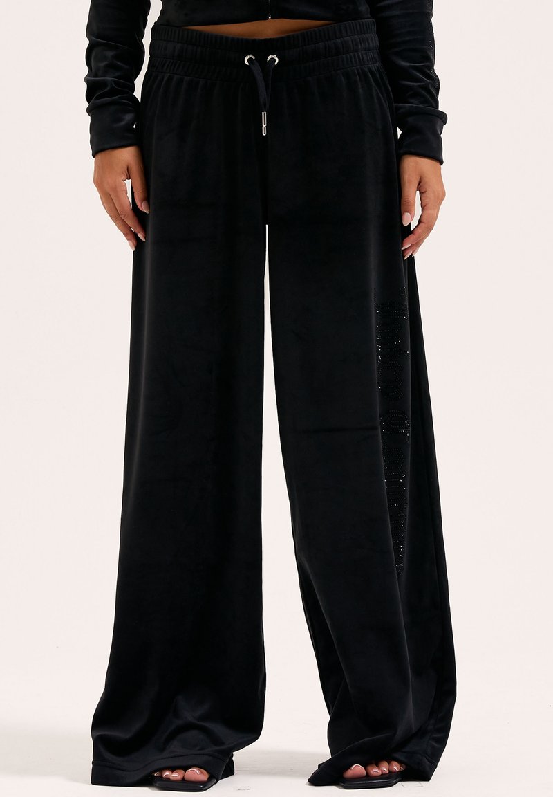 Zwarte wijde pantalon van zacht, fluwelig materiaal. Heeft een elastische tailleband en een zijdetail met kristalversiering.