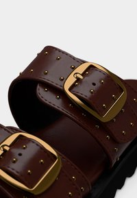 Lanières de sandales en cuir marron avec boucles dorées et petits clous ronds sur une semelle noire, vue rapprochée.