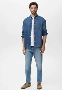 Camicia in denim con bottoni, azzurra, a maniche lunghe e una tasca sul petto. Abbinata a jeans blu sbiaditi e scarpe beige.