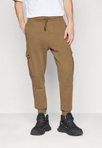 Bruna joggingsbyxor med elastisk midja, snörning, sidofickor och ribbade muddar, i kombination med svarta träningsskor.