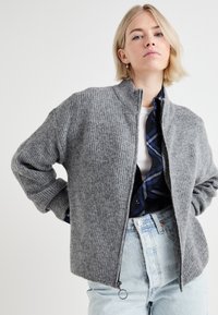 Veste en tricot côtelé gris avec un col haut et une fermeture éclair sur le devant, présentant une coupe décontractée et des épaules tombantes. Portée par-dessus une chemise à carreaux bleue.