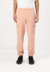 Lys rosa joggebukser med elastisk liv, sidelommer, og ribbede mansjetter, sammen med svarte joggesko. Myk, stretchy stoff.