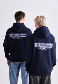 Les sweats à capuche bleu marine présentent un logo imprimé "tommy jeans" avec des noms de villes en dessous. Design décontracté avec cordons de serrage et poignets côtelés.