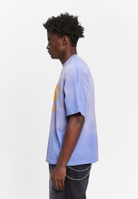 Lys lilla bomulls t-skjorte med korte ermer, prydet med oransje grafisk trykk. Brukt sammen med mørke jeans og en kjedeaccents på lommen.