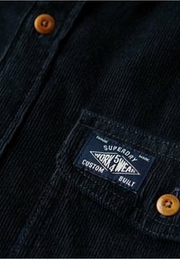 Zwarte corduroy overhemd met een borstzak en een blauw label met de tekst "Superdry Workwear Custom Built," versierd met een bruine knoop.