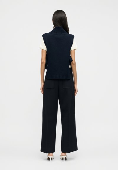 Μπλε σκούρο πλεκτό αμάνικο γιλέκο με ψηλό γιακά, συνδυασμένο με κρεμ τοπ και μαύρο παντελόνι φαρδιάς γραμμής. Λευκή λεπτομέρεια στο παπούτσι.