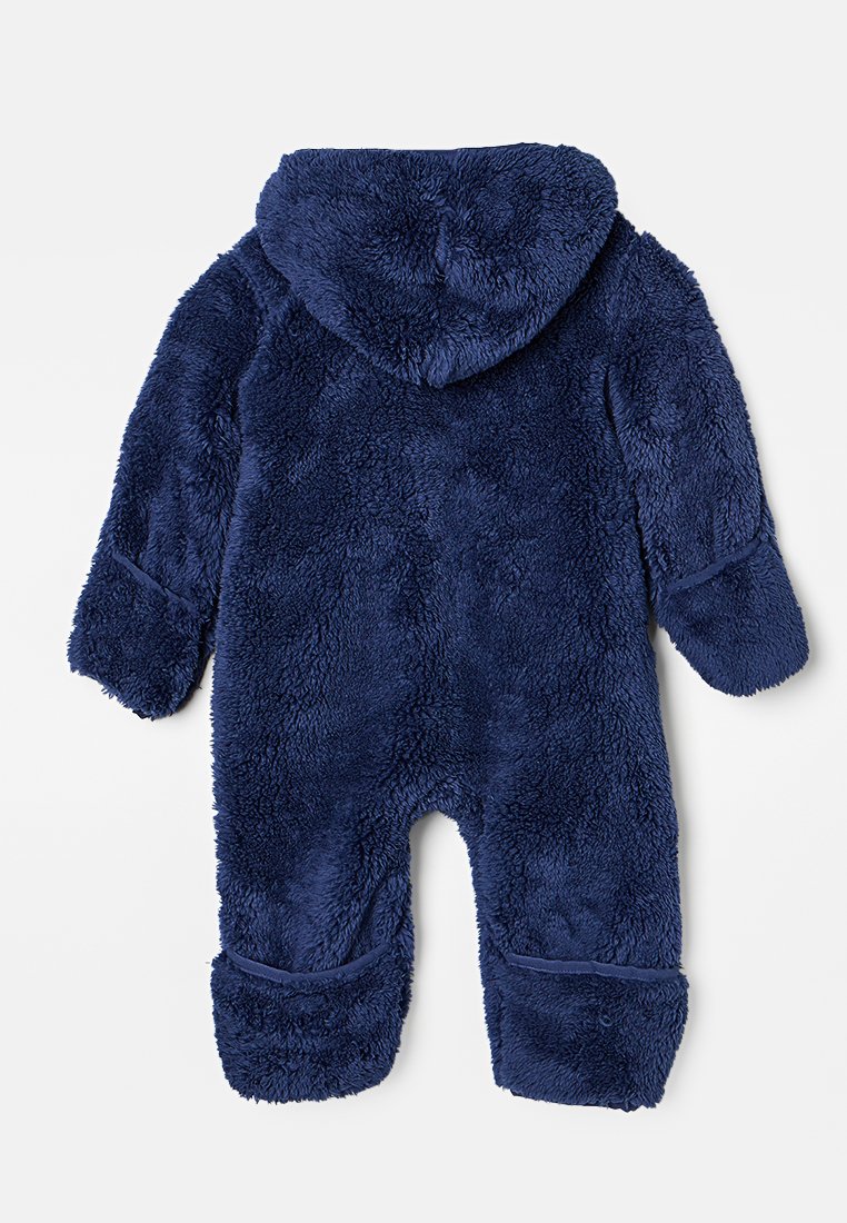Mörkblå fleecepyjamas med huva, långa ärmar och mudd på benen. Mjuk, fluffig textur; sömlös baksida utan synliga knäppningar.