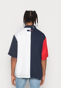 Muž s pletenými vlasy, oblečený v košili s krátkým rukávem, která je rozdělena na vertikální barevné bloky v navy modré, bílé a červené barvě, v kombinaci se světle modrými džíny.