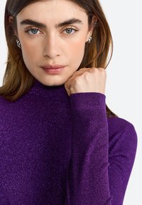 Top lung cu mâneci lungi de culoare purpurie, cu guler înalt, realizat dintr-un material texturat și strălucitor. Pumn strâns la guler, arătând piele netedă și cercei.