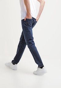 Mörkblå denimjeans med slim fit, som har klassiska sömmar och fem fickor, kombinerat med vita sneakers och en enkel vit t-shirt.