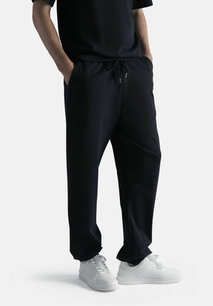 Mies pukeutunut mustiin nyörillisiin housuihin ja valkoisiin tennareihin, kädet taskuissa, seisoo yksinkertaisen vaalean taustan edessä.