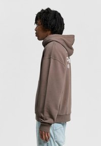 Brown hooded sweatshirt with a front pocket, ribbed cuffs, and hem. Features white text on the back. Soft material with a relaxed fit.